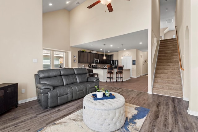 living area with a ceiling fan, stairs, a towering ceiling, and dark wood-style floors
