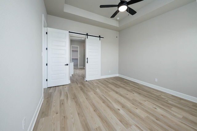 unfurnished bedroom featuring a barn door, a ceiling fan, a tray ceiling, light wood finished floors, and baseboards