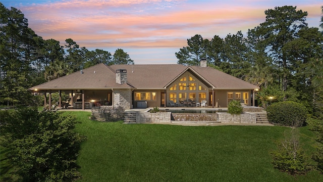 back of house at dusk featuring a chimney, a yard, a tile roof, a patio, and stone siding
