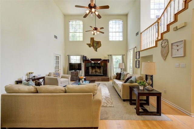 living room featuring a fireplace with raised hearth, a towering ceiling, baseboards, light wood-style flooring, and ceiling fan