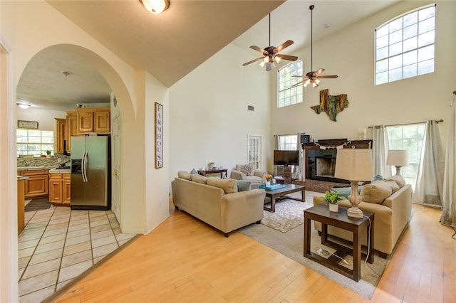 living room featuring a fireplace with raised hearth, light wood-style floors, ceiling fan, arched walkways, and a towering ceiling