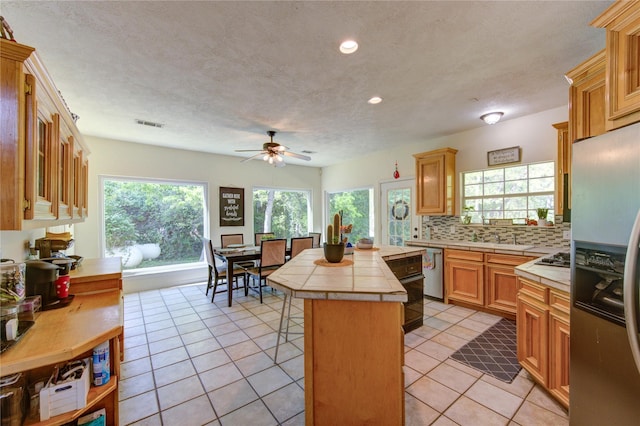kitchen featuring stainless steel appliances, a ceiling fan, tile countertops, light tile patterned floors, and a center island