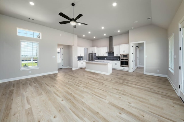 kitchen with open floor plan, stainless steel appliances, wall chimney exhaust hood, a kitchen island with sink, and high vaulted ceiling
