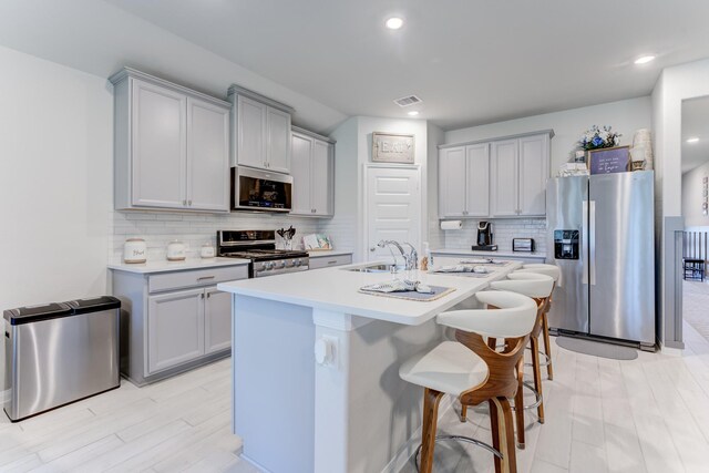 kitchen with stainless steel appliances, a sink, gray cabinets, light countertops, and an island with sink