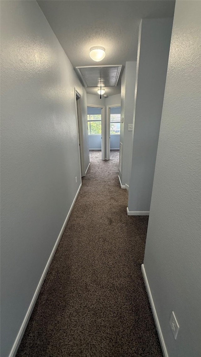 hallway featuring attic access, dark colored carpet, baseboards, and a textured ceiling
