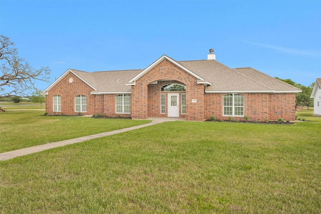 ranch-style house with a chimney, a front yard, a shingled roof, and brick siding