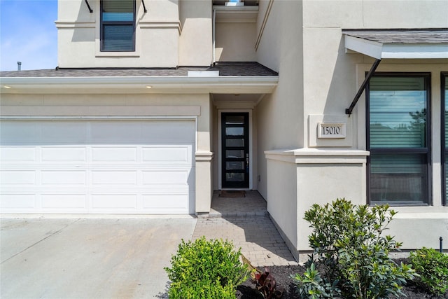 entrance to property featuring a garage, driveway, stucco siding, and roof with shingles