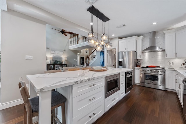 kitchen featuring dark wood-type flooring, a ceiling fan, a kitchen island, wall chimney range hood, and appliances with stainless steel finishes