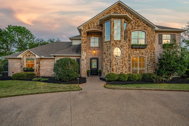 french country home featuring stone siding, a lawn, roof with shingles, and brick siding