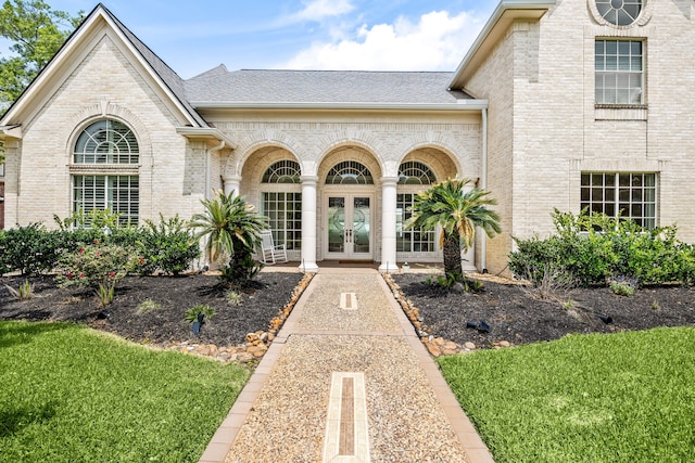 property entrance featuring brick siding, french doors, a lawn, and a shingled roof