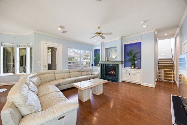 living area with dark wood-type flooring, crown molding, a fireplace, stairs, and ceiling fan