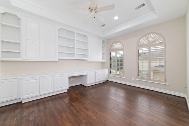unfurnished office with a tray ceiling, dark wood-style flooring, visible vents, built in desk, and ceiling fan