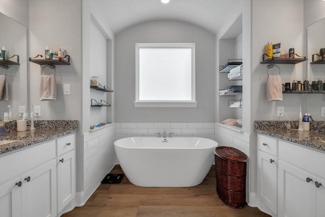 full bath with two vanities, wood finished floors, a freestanding tub, and wainscoting