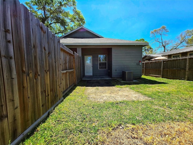 rear view of house with central AC unit, a fenced backyard, a patio area, a lawn, and roof with shingles