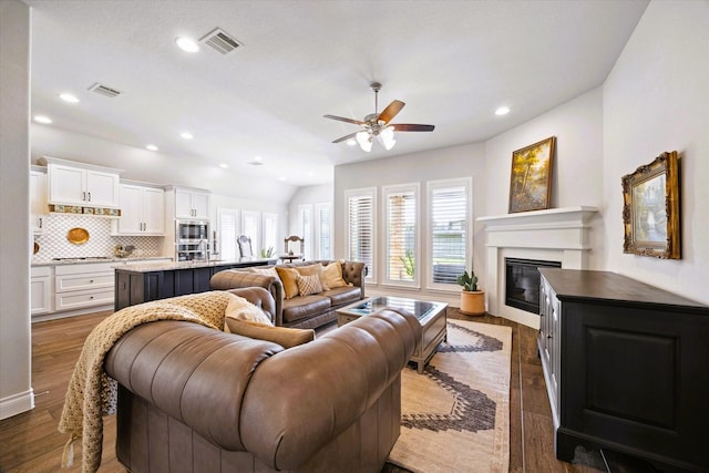 living room featuring ceiling fan, visible vents, dark wood-style floors, and a glass covered fireplace