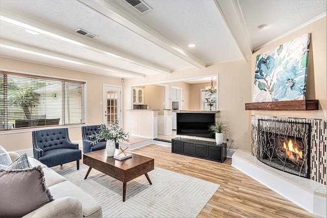 living area featuring a warm lit fireplace, visible vents, beamed ceiling, light wood-style flooring, and a textured ceiling