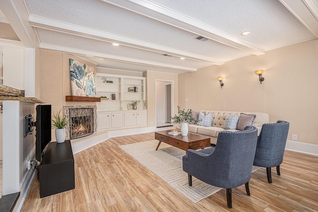 living area with baseboards, a warm lit fireplace, light wood-style flooring, a textured ceiling, and beam ceiling