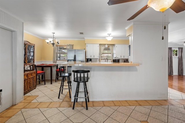 kitchen with a peninsula, stainless steel refrigerator with ice dispenser, ornamental molding, and ceiling fan with notable chandelier