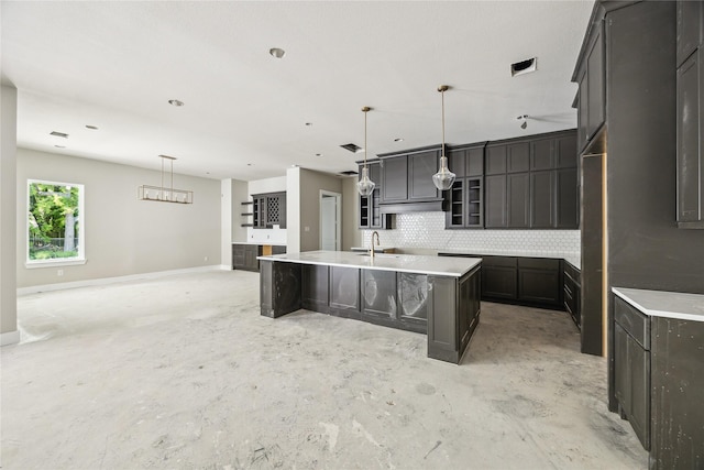 kitchen with a kitchen island with sink, a sink, decorative backsplash, glass insert cabinets, and unfinished concrete floors