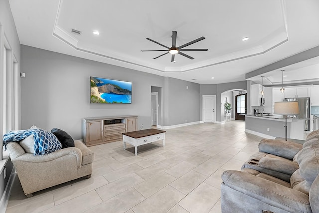 living room with ceiling fan, a raised ceiling, arched walkways, baseboards, and light tile patterned floors