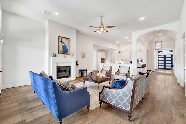 living area with visible vents, arched walkways, light wood-style floors, a glass covered fireplace, and ceiling fan