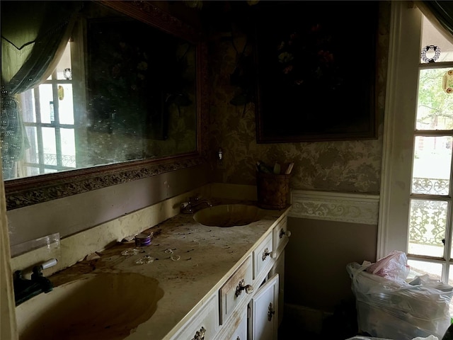 bathroom featuring double vanity, a wainscoted wall, wallpapered walls, and a sink