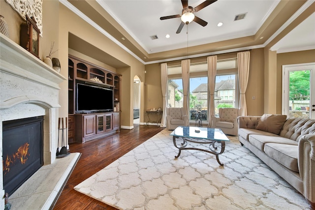 living area featuring a tray ceiling, a premium fireplace, ceiling fan, dark wood finished floors, and ornamental molding