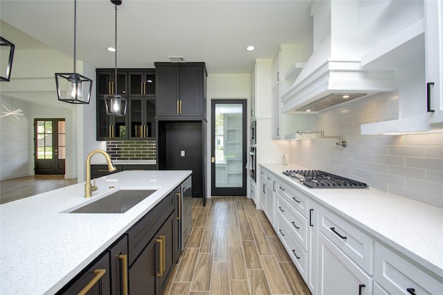 kitchen with white cabinets, backsplash, a sink, premium range hood, and glass insert cabinets