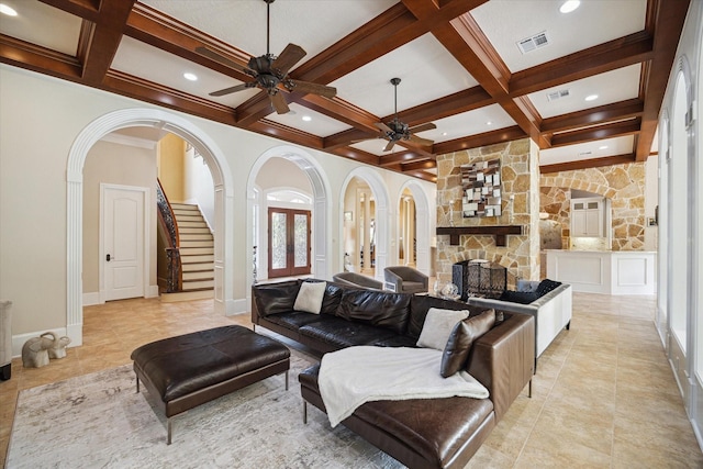 living area with ceiling fan, coffered ceiling, a stone fireplace, visible vents, and arched walkways