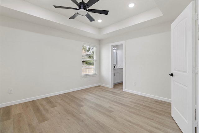 unfurnished bedroom featuring a tray ceiling, light wood-style floors, baseboards, recessed lighting, and ensuite bath