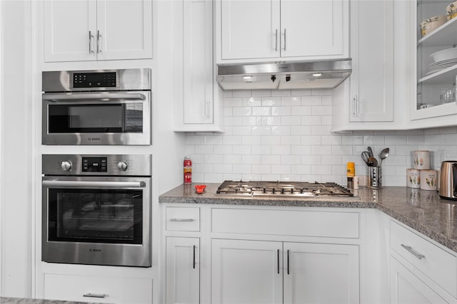 kitchen featuring white cabinets, backsplash, glass insert cabinets, under cabinet range hood, and appliances with stainless steel finishes