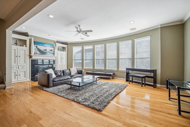 living room with ceiling fan, light wood finished floors, a tiled fireplace, ornamental molding, and visible vents