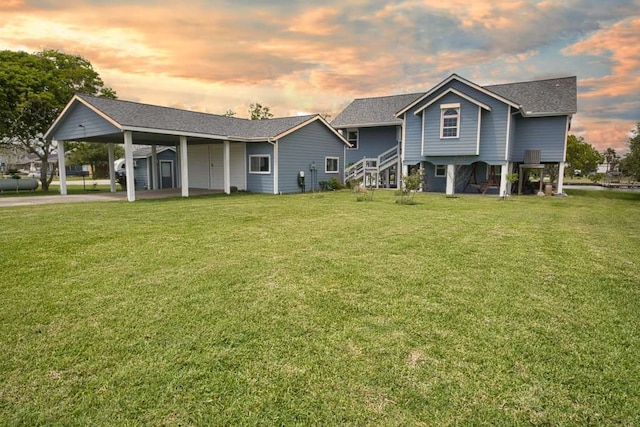 back of house with an attached carport, driveway, a shingled roof, stairway, and a yard
