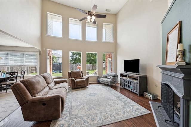 living room featuring visible vents, a towering ceiling, dark wood-type flooring, a fireplace with raised hearth, and a ceiling fan