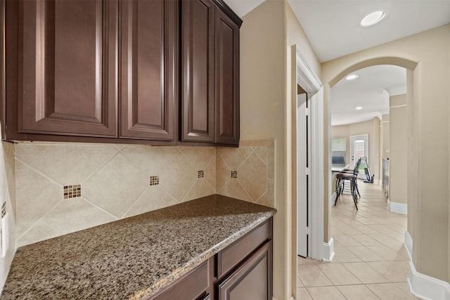 514 Whitney Oaks Ln, Stafford, TX 77477 - kitchen featuring dark stone countertops, dark brown cabinets, light tile patterned floors, arched walkways, and decorative backsplash