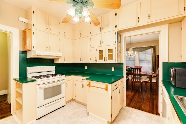 kitchen with under cabinet range hood, white gas range, dark countertops, stainless steel microwave, and ceiling fan with notable chandelier