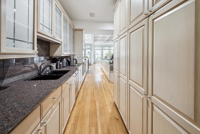 kitchen featuring dark stone countertops, glass insert cabinets, a sink, light wood-type flooring, and backsplash