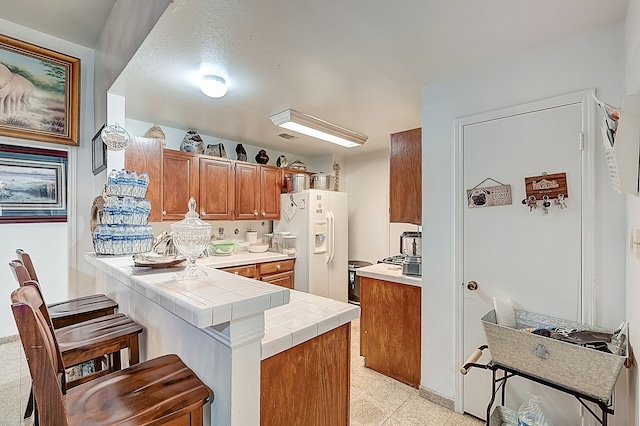 kitchen featuring brown cabinets, tile counters, a peninsula, white fridge with ice dispenser, and a kitchen bar