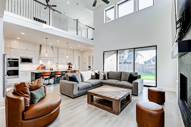 living area featuring a ceiling fan, visible vents, a towering ceiling, a brick fireplace, and light wood-style floors