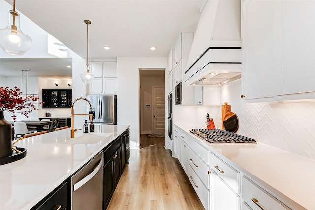 kitchen featuring white cabinets, custom range hood, a sink, light wood-style flooring, and appliances with stainless steel finishes