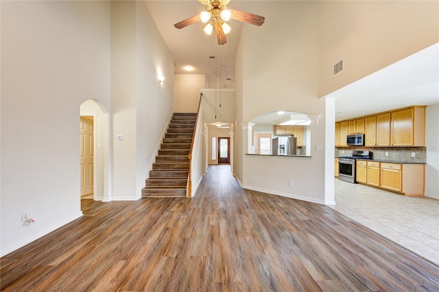 unfurnished living room with ceiling fan, visible vents, wood finished floors, high vaulted ceiling, and stairs