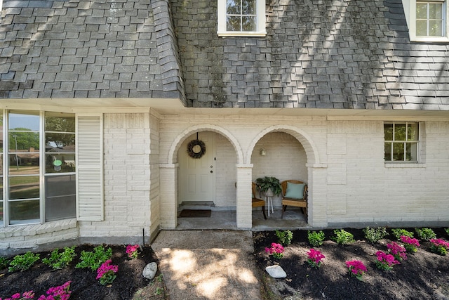 view of exterior entry featuring a porch, brick siding, and roof with shingles