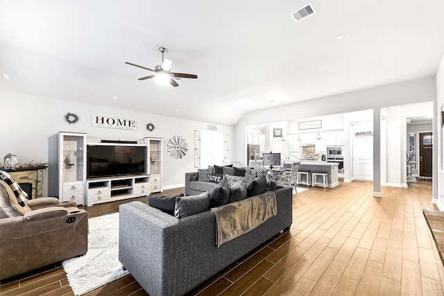 living room featuring vaulted ceiling, a stone fireplace, wood finished floors, ceiling fan, and visible vents