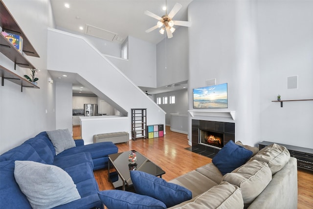living area featuring a ceiling fan, stairs, light wood-style flooring, a tiled fireplace, and a high ceiling