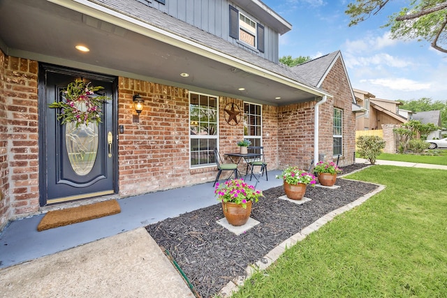 property entrance featuring a lawn, roof with shingles, covered porch, and brick siding
