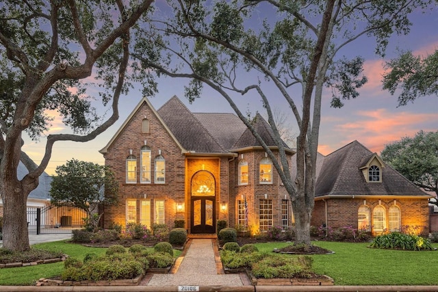 french country inspired facade with roof with shingles, brick siding, and a yard