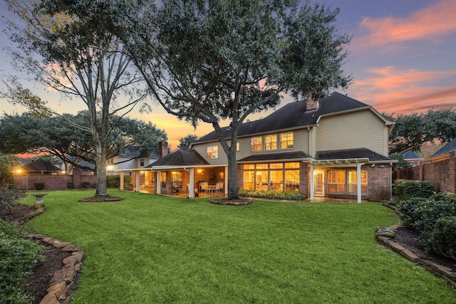 back of property at dusk featuring fence, a chimney, brick siding, and a yard
