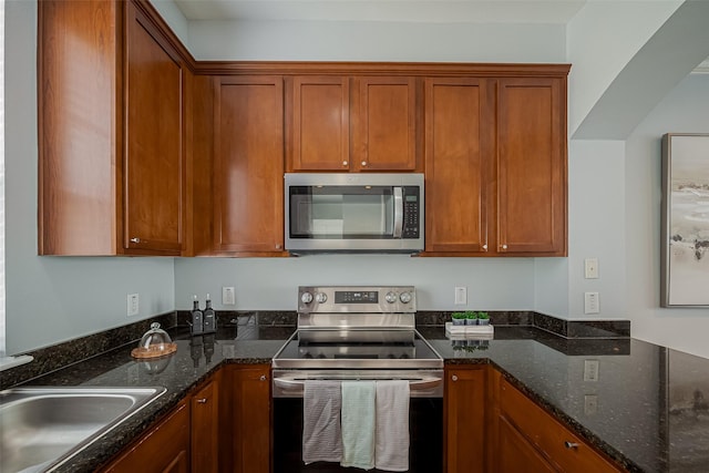 kitchen featuring brown cabinetry, dark stone countertops, stainless steel appliances, and a sink