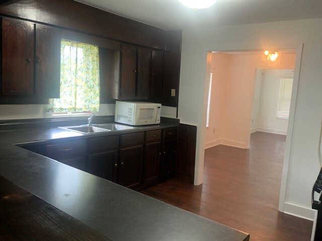 kitchen with dark wood finished floors, dark countertops, a sink, white microwave, and dark brown cabinetry