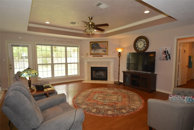 living room with a tiled fireplace, wood finished floors, ornamental molding, visible vents, and a tray ceiling